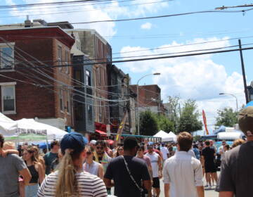 A crowd walks down a street. Buildings are visible in the background.