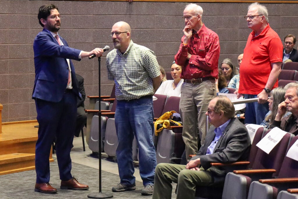 A man is asking a question at a microphone from the audience, with several other people lined up behind him